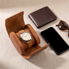 Étui de voyage Escale cognac ouvert avec une montre à cadran crème et bracelet cuir, posé à côté d’un portefeuille, d’un smartphone et de lunettes de soleil, vue de dessus