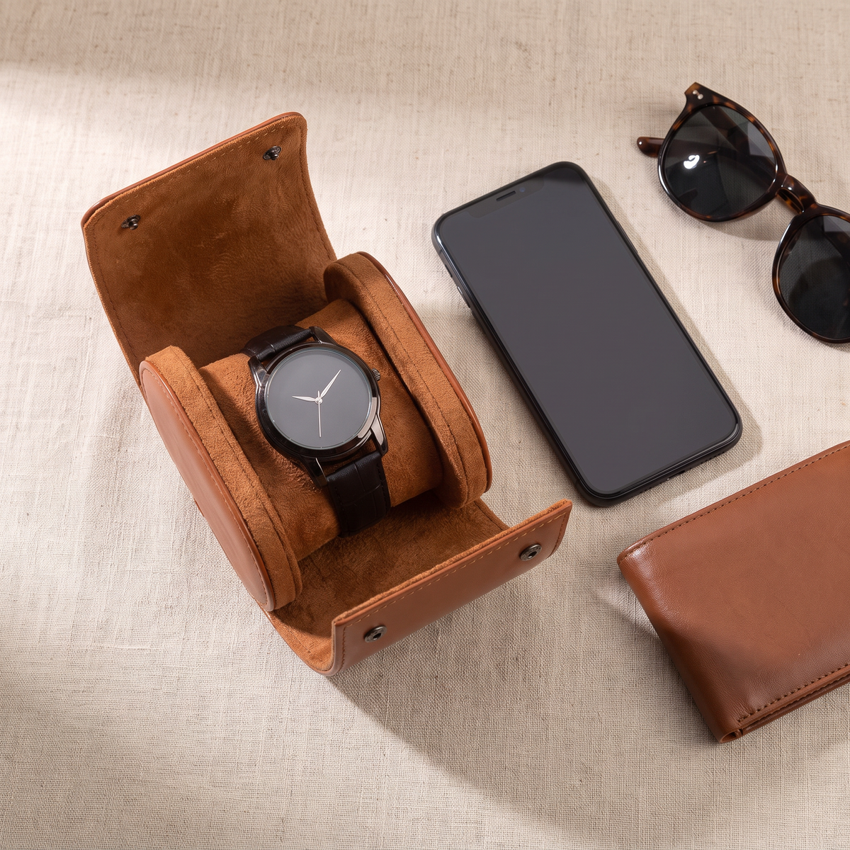 Étui de voyage Escale cognac ouvert avec une montre à cadran noir, posé à côté d’un smartphone, de lunettes de soleil et d’un portefeuille, vue de dessus